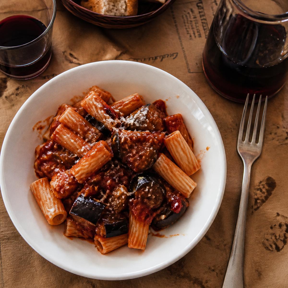 Pasta alle melanzane in a white bowl with a carafe of house red wine